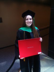 Woman in black and green cap and gown holding a red diploma folder