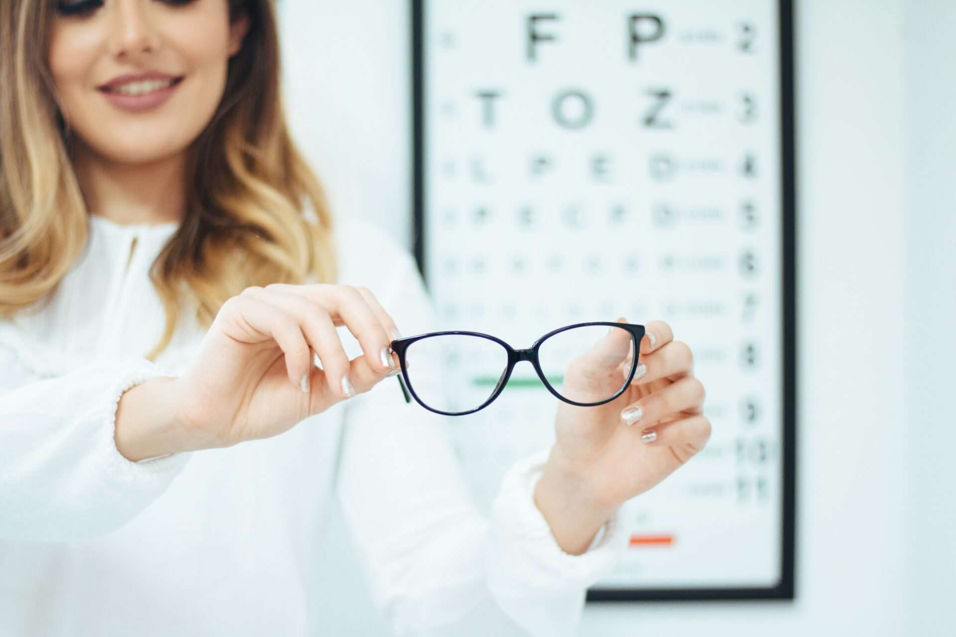 Woman holding a pair of black glasses in front of an eye chart