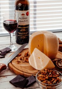 Large wheel of cheese with a wedge cut out. There is a glass of red wine with the bottle. Scattered nuts and chocolate cover the wooden cheese board.